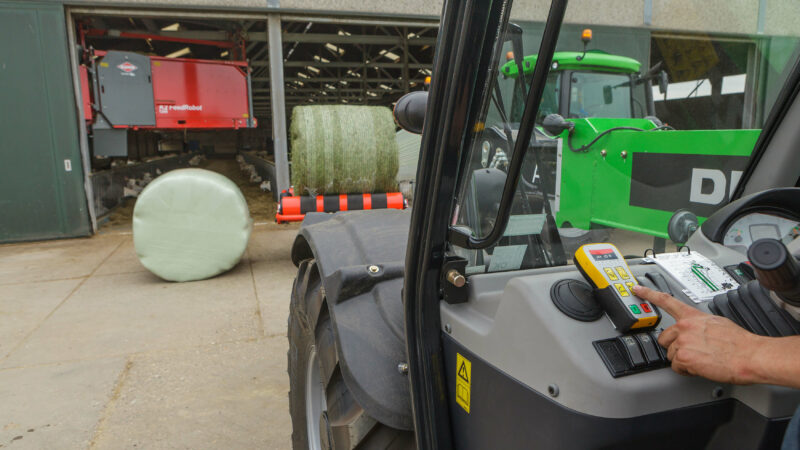 Activation d'un modèle RW 1110 enrubannant une balle ronde devant une étable avec un robot d'alimentation en arrière-plan.