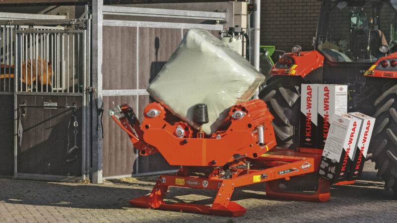 Une enrubanneuse pour balles rectangulaires SW1114 enrubannant une balle en position stationnaire devant une étable.